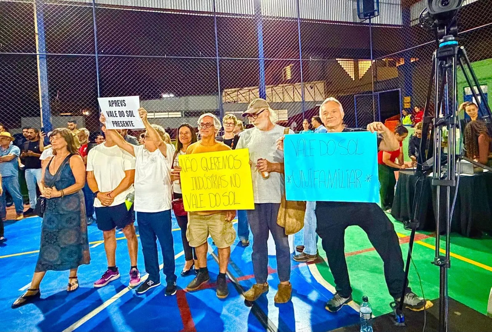 Grupo de moradores numa quadra coberta segurando cartazes com os dizeres "APREVS Vale do Sol presente", "Não queremos indústrias no Vale do Sol" e "Vale do Sol unifamiliar".