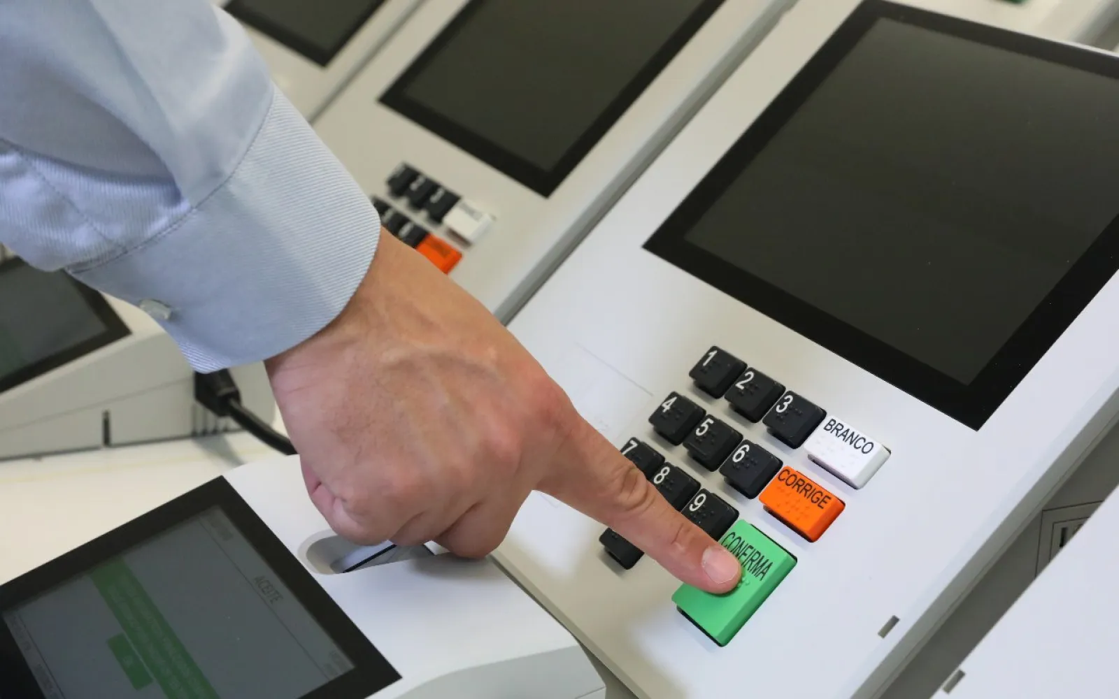 Mão aperta a tecla verde Confirma no teclado de uma urna eletrônica, com outras urnas ao fundo.